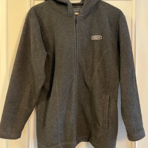 Grå fleecejacka med huva från Chinook - Mysig grå fleecejacka från Chinook Sport med huva och hel dragkedja framtill. Jackan har två sidofickor och en enkel, stilren design. Perfekt för kyliga dagar när du vill vara både bekväm och varm.