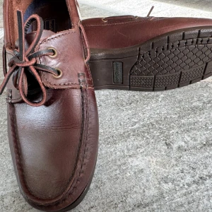 Bruna seglarskor från Henri Lloyd - Klassiska bruna seglarskor från Henri Lloyd i skinn med mörkbruna snören och robust gummisula, funkar året runt. Skorna har metallöljetter och klassisk Union Jack-detalj på sidan. 