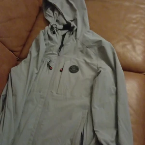 Grå vindjacka från Zavetti Canada - Snygg grå vindjacka från Zavetti Canada med huva och svarta detaljer. Jackan har dragkedja framtill, justerbar huva med snörning och en tydlig logga på bröstet samt tryck på ärmen. Perfekt för blåsiga dagar och streetwear-stil.