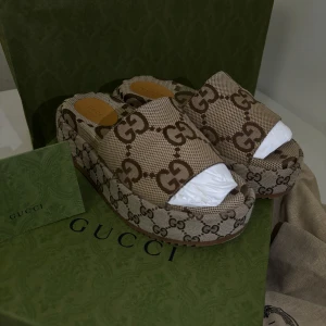 Gucci sandaler med GG-monogram - Snygga sandaler från Gucci med breda remmar och platåsula, täckta i det klassiska GG-monogrammet i beige och brunt. Skorna har öppen tå och är tillverkade i canvas med läderdetaljer på innersulan. Perfekta för dig som vill sticka ut med lyxig vibe.