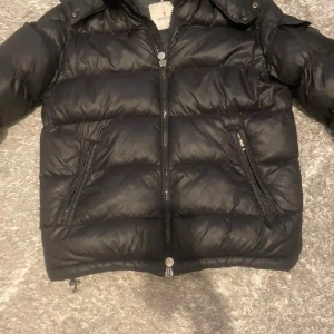 Svart Moncler pufferjacka med dragkedja - Snygg svart pufferjacka från Moncler med klassisk logga på ärmen och tvåvägsdragkedja framtill. Jackan har två snedställda fickor med dragkedja och en glansig finish. Perfekt för kalla dagar och riktigt stilren look.