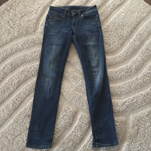 G-Star RAW Midge Mid Straight jeans - Snygga mörkblå jeans från G-Star RAW, modell Midge Saddle Mid Straight. Klassisk femficksdesign, raka ben och midwaist. Jeansen har slitningar och kontrastsömmar för en cool look. Perfekta till sneakers eller boots. Storlek W27 L30 