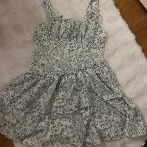 Grön Blommig kortklänning från SHEIN storlek M - Supersöt vit kortklänning från SHEIN med grönt blommönster, volangig kjol och breda axelband med knyt. Tillverkad i mjuk polyesterblandning med lite stretch. Perfekt för sommarens alla tillfällen när du vill känna dig extra fin.