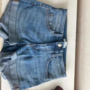 Blå jeansshorts med upprullad kant - Snygga blå jeansshorts i klassisk modell med upprullad kant och femficksdesign. Shortsen har bälteshällor, knapp och dragkedja framtill samt bakfickor. Perfekta för varma dagar och enkel att matcha med allt.