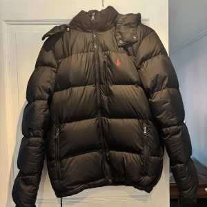 Svart pufferjacka från Polo Ralph Lauren - Svart pufferjacka från Polo Ralph Lauren med röd broderad logga på bröstet. Jackan har hög krage, avtagbar huva och flera dragkedjeförsedda fickor. Perfekt för kalla dagar med sin vadderade design och klassiska look. Den har även ett band längst ner ifall man vill ha den lite tajtare, knappt andvänd. Priset kan diskuteras, köparen står för frakt!