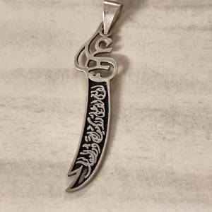 Silvrigt halsband med svärdberlock - Unikt halsband med en svärdformad berlock i silverfärgad metall. Ena sidan har svart bakgrund med detaljerat mönster och inskription, medan andra sidan är slät. Berlocken har en cool, böjd form och ett dekorativt övre parti.