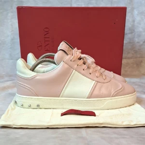 Valentino Flycrews sneakers rosa/vit - Snygga Valentino Garavani Open sneakers i ljusrosa och vitt skinn. Klassisk låg modell med snörning, rund tå och platt sula. Ikoniska nitar i silver på hälen ger extra edge. Perfekta för dig som vill ha en lyxig och stilren sneaker med exklusiv känsla.