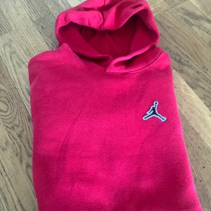 Röd Jordan hoodie med logga - Snygg röd hoodie från Jordan med klassisk Jumpman-logga på bröstet. Tröjan har huva och en stor magficka framtill. Perfekt för dig som gillar streetwear och sportig stil. Tillverkad i mjuk bomullsmix för skön känsla hela dagen.