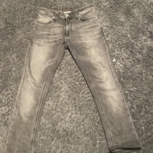 Grå skinny jeans från Nudie Jeans - Snygga grå jeans från Nudie Jeans i slim fit. Jeansen har klassisk femficksdesign, svarta vågdetaljer på bakfickorna och en tvättad look. Perfekta för dig som gillar en smal siluett och vill ha ett par stilrena jeans i garderoben. Storlek L30 W25 men de har krympt i tvätten så skulle säga mer L29-L28