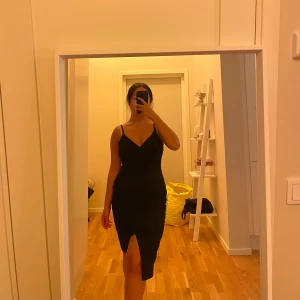 Svart klänning med slits och smala axelband - Säljer en svart, figurnära klänning med smala axelband och v-ringning. Klänningen har en snygg slits framtill och är midilång, perfekt för dig som gillar en stilren och elegant look. Materialet känns mjukt och följsamt mot kroppen.