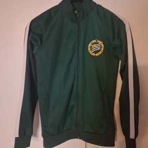 Grön Hammarby ziptröja XS - Grön äldre modell ziptröja från Hammarby Officiell med klubbens retro emblem broderat på bröstet. Tröjan har vita ränder längs ärmarna, hög krage och två fickor framtill. Tillverkad i polyester och perfekt för dig som vill visa ditt stöd på matchen eller i vardagen. Forza bajen!