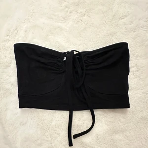 Svart bandeau tubtopp från Noisy May - Svart bandeau tubtopp från Noisy May med snygg snörning framtill och knytband som kan bäras runt nacken. Toppens tyg är mjukt och stretchigt, perfekt till sommarens outfits. Enkel och stilren design som är lätt att matcha.