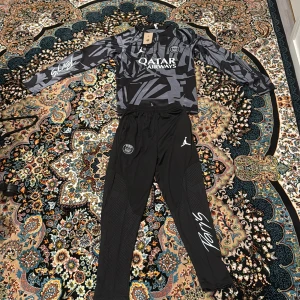 PSG långärmad fotbollströja Jordan - Snygg långärmad PSG fotbollströja från Jordan med svart och grått grafiskt mönster, dragkedja vid halsen och Qatar Airways-tryck på bröstet. Tillverkad i lätt och ventilerande material, perfekt för träning eller match.