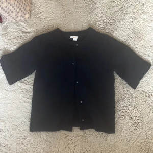 Svart kofta från Gina Tricot - Mysig marin blå kofta från Gina Tricot med kort ärm och rund hals. Koftan är stickad och har knappar framtill i matchande färg. Perfekt för lager-på-lager och enkel att styla till olika outfits.