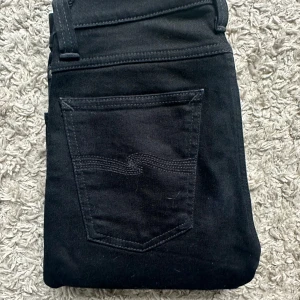 Svarta nudie jeans - Svarta jeans med smal passform och klassisk femficksdesign. Byxorna har en enkel bakficka med diskret söm och är tillverkade i stretchigt jeanstyg för en tight och bekväm känsla. Perfekta för dig som gillar en stilren och slimmad look. 27/32