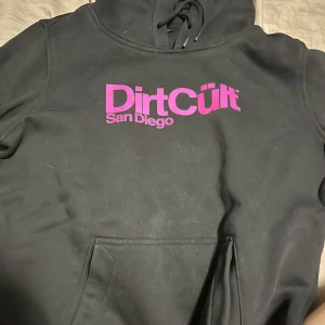 Svart hoodie DirtCült San Diego - Svart hoodie från DirtCült med stor magficka och huva med snörning. Tryck i stark rosa färg på bröstet med texten 'DirtCült San Diego'. Skön och casual passform, perfekt till chill dagar. Materialet känns mjukt och är troligtvis bomull eller bomullsblandning. Lite hårig men det går bort