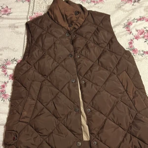 Brun quiltad dunväst från Freequent - Quiltad brun dunväst från Freequent i storlek XS. Västen har hög krage, tryckknappar framtill och två fickor. Perfekt för lager-på-lager och ger en snygg, avslappnad look. Fodret är beige och materialet känns lätt och smidigt.