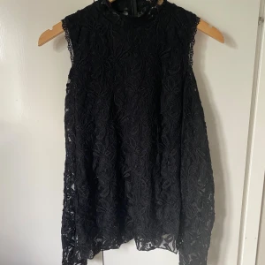 Svart spetstop från Amisu - Supersnygg svart kortklänning från Amisu i spets med blommigt mönster. Hög hals, ärmlös design och dragkedja bak. Perfekt för dig som vill ha en elegant look med lite edge. Klänningen har en loose passform och är helfodrad.