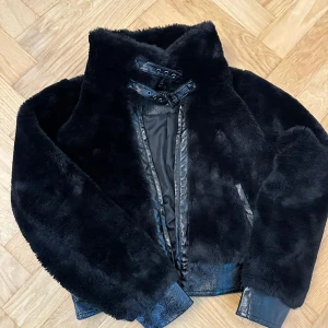 Svart pälsjacka med läderdetaljer - Säljer en svart pälsjacka från NA-KD med snygga läderdetaljer vid krage och ärmslut. Jackan har hög krage med spännen och är perfekt för kalla dagar. Den är fluffig och har en cool, modern look.