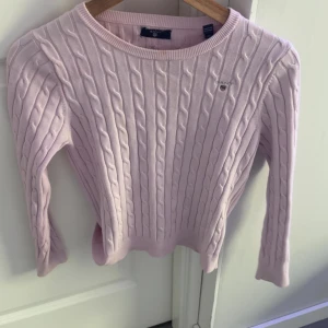 Ljusrosa kabelstickad tröja från Gant - Supersöt ljusrosa kabelstickad tröja från Gant med rund halsringning och diskret logga på bröstet. Tröjan har lång ärm och klassisk ribbad kant vid hals, ärmslut och nederkant. Perfekt för dig som gillar preppy och stilren look.