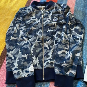 NBA Kings camo ziptröja blå/grå - Säljer en blå och grå camouflagemönstrad ziptröja från NBA Kings. Tröjan har hel dragkedja, ribbade muddar och en broderad Kings-logga på bröstet. Perfekt för dig som gillar basket och streetwear.