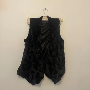Svart pälsväst från Boutique L - Mysig svart pälsväst från Boutique i storlek L. Västen har en mjuk, fluffig yta och är helsvart med lätt glansigt innerfoder. Modellen är öppen framtill och har en avslappnad passform med något längre framstycken. Har även två fickor. 