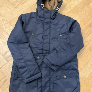 Mörkblå parkajacka med huva - Snygg mörkblå parkajacka med huva och dragsko, perfekt för kyliga dagar. Jackan har flera fickor med knappar och dragkedja framtill. Fodrad insida i kontrastfärg och långa ärmar. Stilren och praktisk design som håller dig varm.