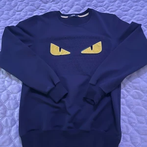 Fendi svart sweatshirt med monsterögon - Säljer en svart sweatshirt från Fendi med ikoniska monsterögon i gult framtill. Tröjan har rund hals, ribbade muddar och diskret Fendi-mönster i tyget. Perfekt för dig som vill sticka ut med en lyxig och edgy vibe.