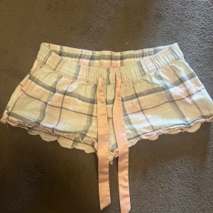 Rutiga shorts med snörning från Hunkemöller - Säljer ett par rutiga shorts från Hunkemöller i ljusa toner av beige, rosa och grått. De har en bred resår i midjan, dekorativ snörning framtill i satin och söt vågig kant nedtill. Perfekta för chill dagar hemma.