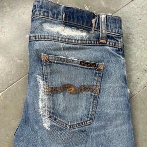 Blå slitna jeans från Nudie Jeans - Säljer ett par blå jeans från Nudie Jeans med tydliga slitningar och lagningar för en riktigt rå streetlook. Klassisk femficksmodell med orange sömmar och Nudie-logga på bakfickan. Jeansen är i denim och har en rak passform.