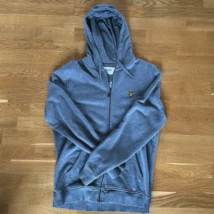 Grå hoodie från Lyle & Scott - Grå hoodie från Lyle & Scott med dragkedja och huva. Tröjan har en gul broderad logga på bröstet, ribbade muddar och fickor framtill. Tillverkad i mjuk bomull, perfekt för chill dagar eller när du vill ha en clean look.