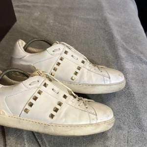 Valentino Rockstud vita sneakers - Snygga vita Valentino Rockstud sneakers i skinn med ikoniska guldiga nitar längs sidorna och över tungan. Klassisk rund tå, platt sula och snörning framtill. Perfekta för dig som vill ha en lyxig och edgy touch till din outfit.