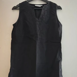 Svart broderad linnetopp från Cappuccini - Svart elegant ärmlös linnetopp från Cappuccini med snygga broderade detaljer framtill och längs nederkanten. Toppen har v-ringning och små slitsar i sidorna. Perfekt för dig som gillar stilrena plagg med unika detaljer.