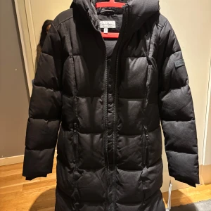 Svart lång pufferjacka Calvin Klein M - Svart lång pufferjacka från Calvin Klein med huva och dragkedja framtill. Jackan har quiltad design, två fickor med dragkedja och mjukt foder i huvan som get mjuk känsla. Perfekt för kalla vinter dagar.🥰Kontakta för fler bilder eller frågor!