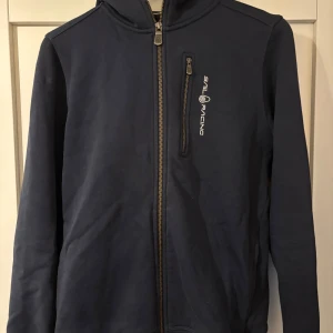 Mörkblå hoodie från Sail Racing - Mörkblå hoodie från Sail Racing med dragkedja framtill och logga på bröstet. Tröjan har huva med broderad detalj och en praktisk bröstficka med dragkedja. Perfekt för dig som gillar sportig och stilren look.
