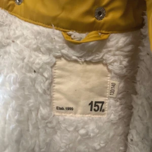 Gul teddyjacka med foder från 157 - Gul teddyjacka från 157 med vit fluffig insida och huva. Jackan har tryckknappar i silver och stora fickor framtill. Perfekt för kalla dagar med sitt mysiga teddyfoder och snygga streetstil.