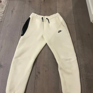 Beige mjukisbyxor från Nike med snörning - Snygga beige mjukisbyxor från Nike med svart logga på låret och svart snörning i midjan. Byxorna har en svart ficka med dragkedja på sidan och är tillverkade i mjukt bomullsmaterial. Perfekta för chill eller träning.