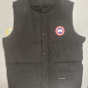 Svart dunväst från Canada Goose - Svart dunväst från Canada Goose med klassisk logga på bröstet och diskret etikett på fickan. Västen har hög krage, tryckknappar framtill och två stora fickor. Perfekt för lager-på-lager och riktigt snygg till streetwear.