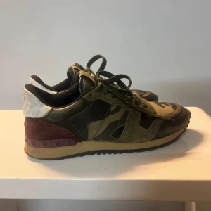 Valentino Rockrunner sneakers camo - Säljer ett par Valentino Rockrunner sneakers i camo-mönster med gröna, svarta och vinröda detaljer. Skorna har snörning, platt sula och coola nitar på hälen. Materialmix av mocka, skinn och textil. Perfekta för dig som vill sticka ut med en lyxig streetstyle.