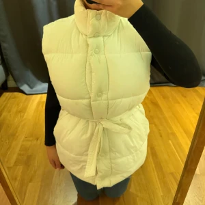 Vit dunväst med bälte från Bik Bok - Säljer en fluffig och quiltad vit dunväst från Bik Bok i storlek S. Västen har hög krage, knappar framtill och ett snyggt knytbälte i midjan som ger en schysst siluett. Perfekt att slänga över en hoodie eller långärmad tröja när det är kyligt ute.