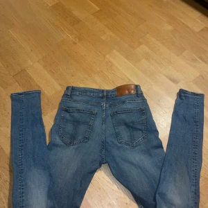 Blå jeans från Tiger of Sweden - Snygga blå jeans från Tiger of Sweden med klassisk femficksdesign och läderpatch bak i midjan. Jeansen har smal passform och raka ben, tillverkade i mjukt denimtyg. Perfekta för dig som gillar stilrena och tidlösa jeans.