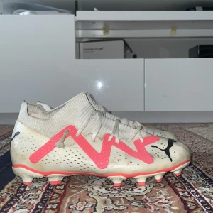 Puma future fotbollsskor  - Säljer ett par snygga fotbollsskor från Puma i vitt med rosa detaljer och svarta loggor. Skorna har snörning, stickad strumpkrage och platt sula med dobbar för bra grepp på planen. Perfekta för dig som vill sticka ut på matchen!