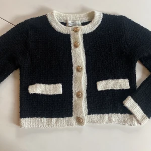 Svartvit kofta från H&M med guldknappar - Snygg svart kofta från H&M med vita detaljer längs kanter, fickor och ärmslut. Koftan har fyra dekorativa guldknappar framtill och är stickad i ett mjukt material. Perfekt för dig som gillar klassisk men trendig stil.