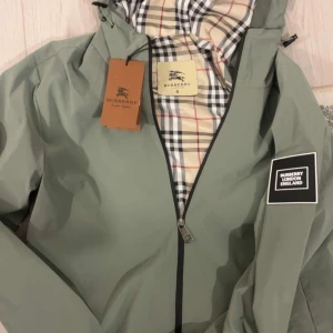 Grön vindjacka från Burberry - Snygg grön vindjacka från Burberry med klassiskt rutigt foder och svartvit logotyp-patch på ärmen. Jackan har dragkedja framtill och huva, perfekt för dig som vill ha en stilren och ikonisk look.