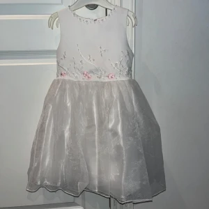 Vit kort klänning med blommor och rosett - Superfin vit kort barnklänning med glansig kjol och broderade blommor i rosa och vitt på livet. Klänningen har ärmlös design, rund halsringning och en söt rosett baktill i midjan. Perfekt för speciella tillfällen när du vill känna dig extra fin.