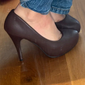 Bruna pumps från Elissa med klack - Säljer ett par stilrena bruna pumps från Elissa med hög klack och rund tå. Skorna har en slät yta och är tillverkade i syntetmaterial. Perfekta för dig som vill ha en klassisk och elegant look.