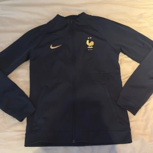 Nike Frankrike ziptröja marinblå S - Marinblå ziptröja från Nike med Frankrikes landslagsemblem och guldiga detaljer. Slim fit-modell med hel dragkedja, långa ärmar och FRANCE-tryck på ryggen. Perfekt för fotbollsfans som vill ha en sportig och snygg look.