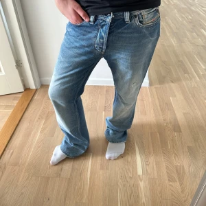 Blå Diesel Rayan jeans bootcut - Säljer ett par blå Diesel Rayan jeans med bootcut passform. Jeansen har slitna detaljer och ljusare partier på benen för en cool, vintage vibe. Klassisk femficksmodell med knappgylf och robust denimkänsla. Perfekt för dig som gillar avslappnad stil.