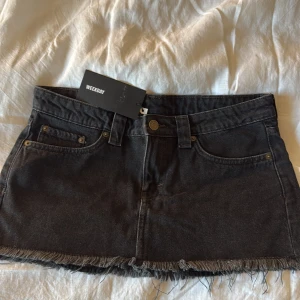 Svart jeanskjol från Weekday, strl 36 - Snygg svart jeanskjol från Weekday i klassisk femficksmodell. Kjolen är kort med rå, fransig kant nertill och har bälteshällor samt knapp och dragkedja framtill. Perfekt för dig som gillar en edgy och avslappnad stil. Den är helt ny och aldrig använt!