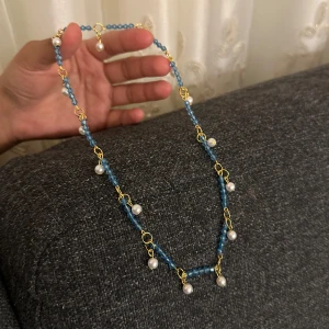 Halsband med blå pärlor och gulddetaljer - Unikt halsband med små blå pärlor och guldfärgade länkar, dekorerat med vita pärlhängen. Halsbandet har en elegant och lekfull design som verkligen sticker ut. Perfekt för dig som vill ha något färgglatt och trendigt i smyckesamlingen.